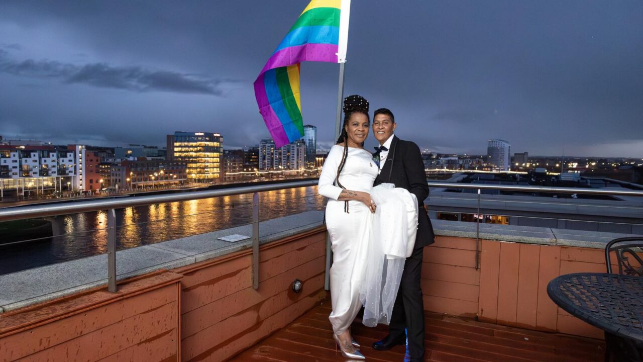 Two people in formal attire stand on a rooftop in the evening, holding a rainbow pride flag, with a cityscape and river in the background.