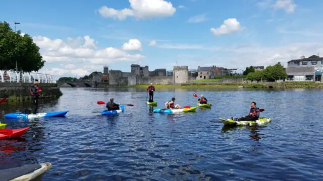 People kayaking on a river near a stone bridge and historic castle under a partly cloudy sky. Buildings and trees line the riverbank in the background.