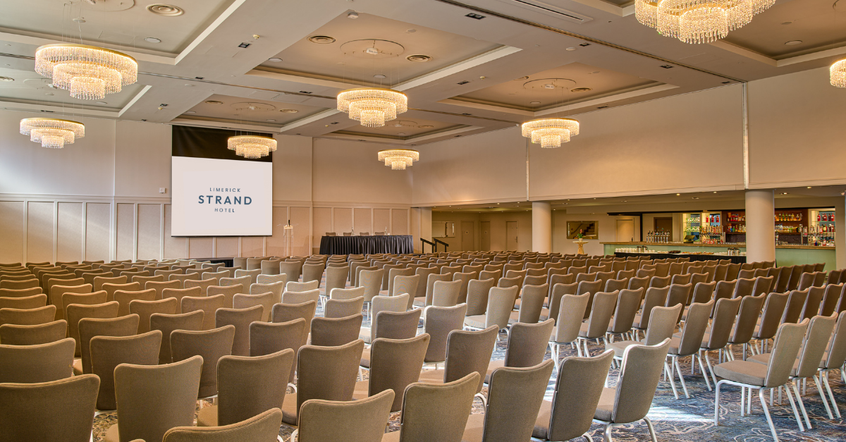 A conference room with rows of empty chairs facing a stage and large screen, featuring modern chandeliers and a bar area on the right.