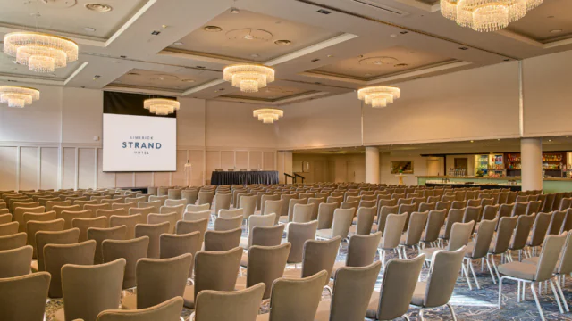 A conference room with rows of empty chairs facing a stage and large screen, featuring modern chandeliers and a bar area on the right.