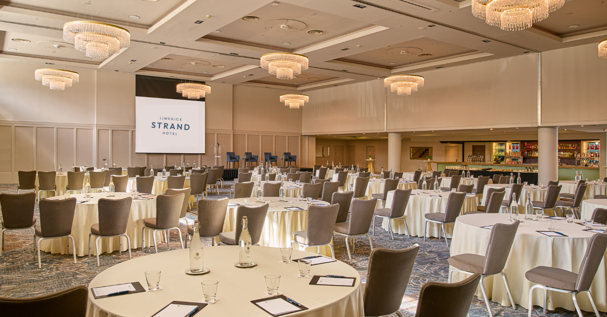 Large hotel conference room with round tables set with notepads, pens, and water bottles, facing a projector screen and stage; chandeliers hang from the ceiling.