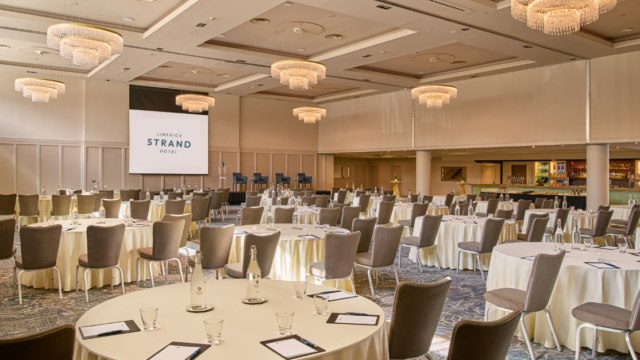 Large hotel conference room with round tables set with notepads, pens, and water bottles, facing a projector screen and stage; chandeliers hang from the ceiling.