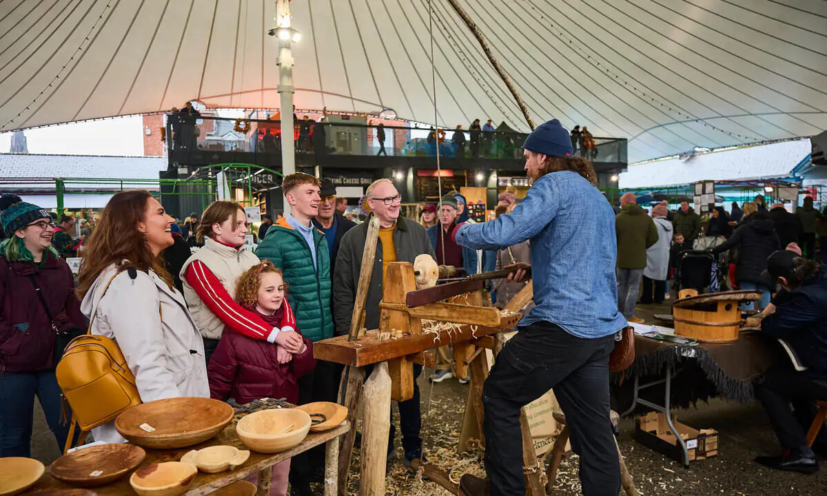 A man demonstrates wood carving to a group of onlookers under a large tent at a busy market or fair, with handmade wooden bowls displayed on tables.