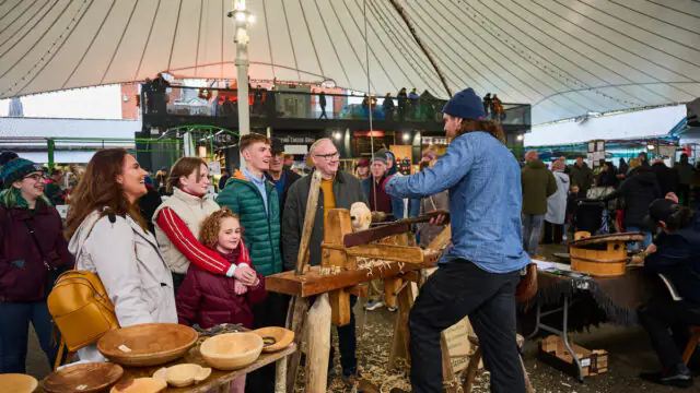 A man demonstrates wood carving to a group of onlookers under a large tent at a busy market or fair, with handmade wooden bowls displayed on tables.