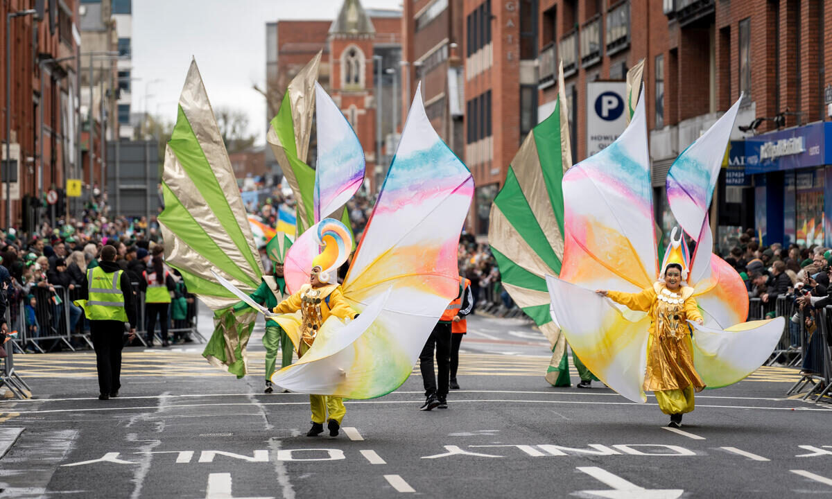 Two parade performers in colorful butterfly costumes walk down a city street, watched by crowds behind barriers during a public event.