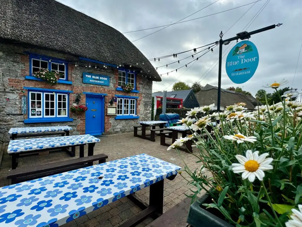 Outdoor seating area with blue patterned tables in front of The Blue Door Restaurant, a stone building with a thatched roof and flowers, under a cloudy sky.
