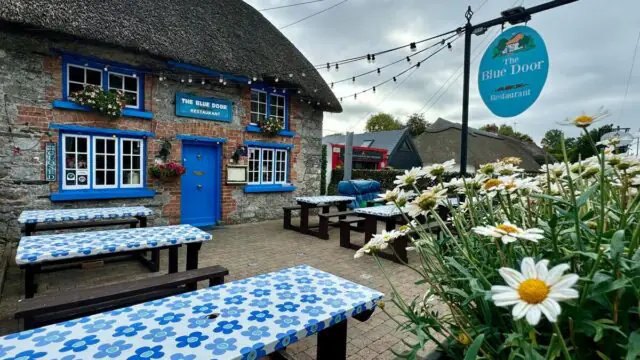 Outdoor seating area with blue patterned tables in front of The Blue Door Restaurant, a stone building with a thatched roof and flowers, under a cloudy sky.