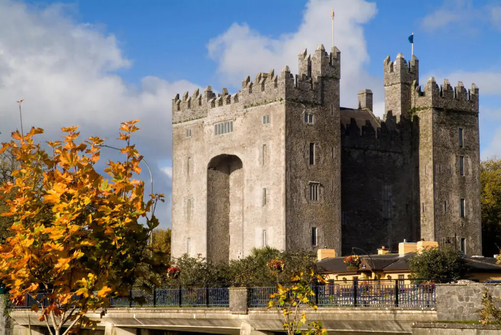 A large stone castle with crenellated towers stands behind trees with autumn foliage under a partly cloudy sky.