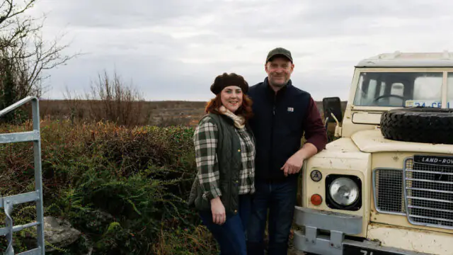 Two people stand outdoors next to a vintage cream-colored Land Rover with rural landscape and cloudy sky in the background.