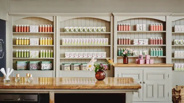 Shelves display neatly arranged bottles, boxes, and jars of personal care or beauty products in a well-lit, tidy shop interior with a wooden countertop and decorative flowers.
