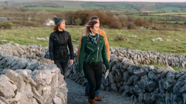 Three people walk along a stone path bordered by low stone walls, with green fields and distant hills in the background.