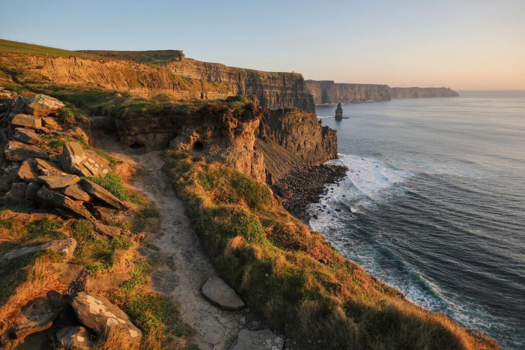 A rugged coastal path runs along the grassy cliffs overlooking the ocean at the Cliffs of Moher in Ireland during sunset.