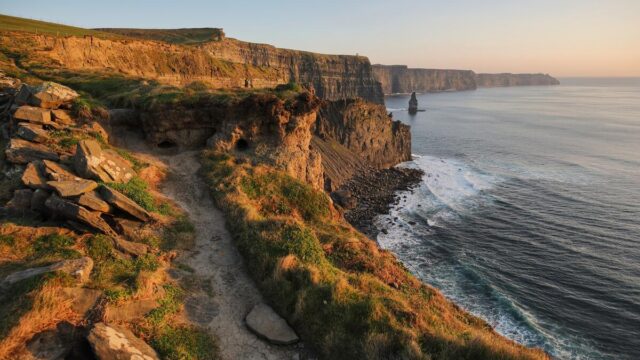 A rugged coastal path runs along the grassy cliffs overlooking the ocean at the Cliffs of Moher in Ireland during sunset.
