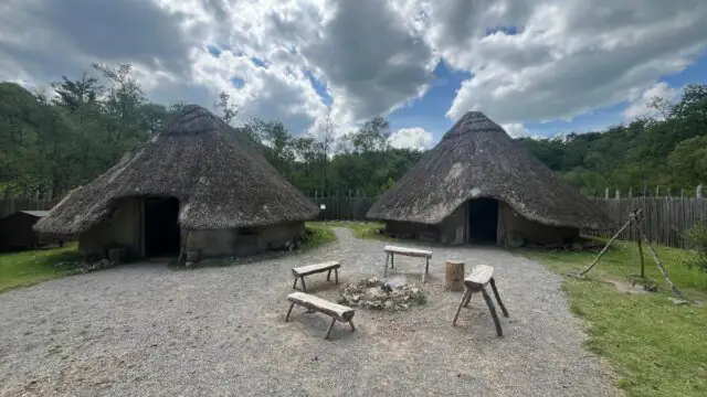 Two round, thatched-roof huts stand in a clearing, with benches, a fire pit, and wooden tools arranged in the foreground under a partly cloudy sky.