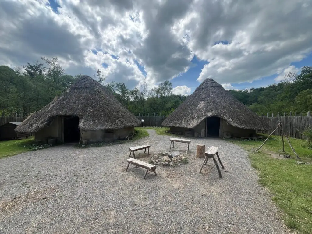 Two round, thatched-roof huts stand side by side, surrounded by trees and a wooden fence, with benches and a fire pit in the foreground under a cloudy sky.
