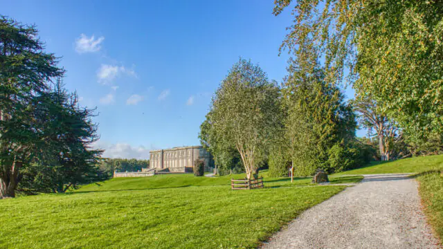 A gravel path curves through a grassy parkland toward a large, rectangular stone building under a clear blue sky.