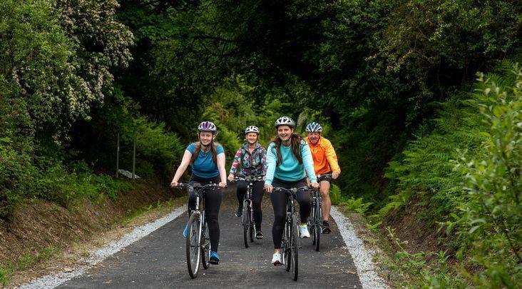 Four people ride bicycles on a paved path surrounded by lush green trees and plants, all wearing helmets and casual clothing.