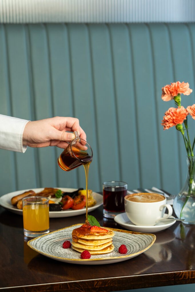 A hand pours syrup onto a stack of pancakes garnished with strawberries and raspberries. Breakfast drinks and food are on the table, with a vase of flowers in the background.