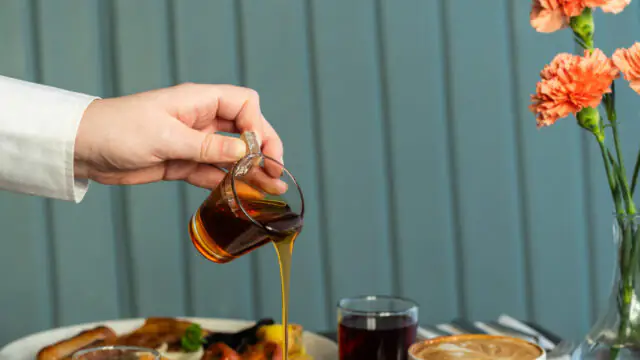 A hand pours syrup onto a stack of pancakes garnished with strawberries and raspberries. Breakfast drinks and food are on the table, with a vase of flowers in the background.