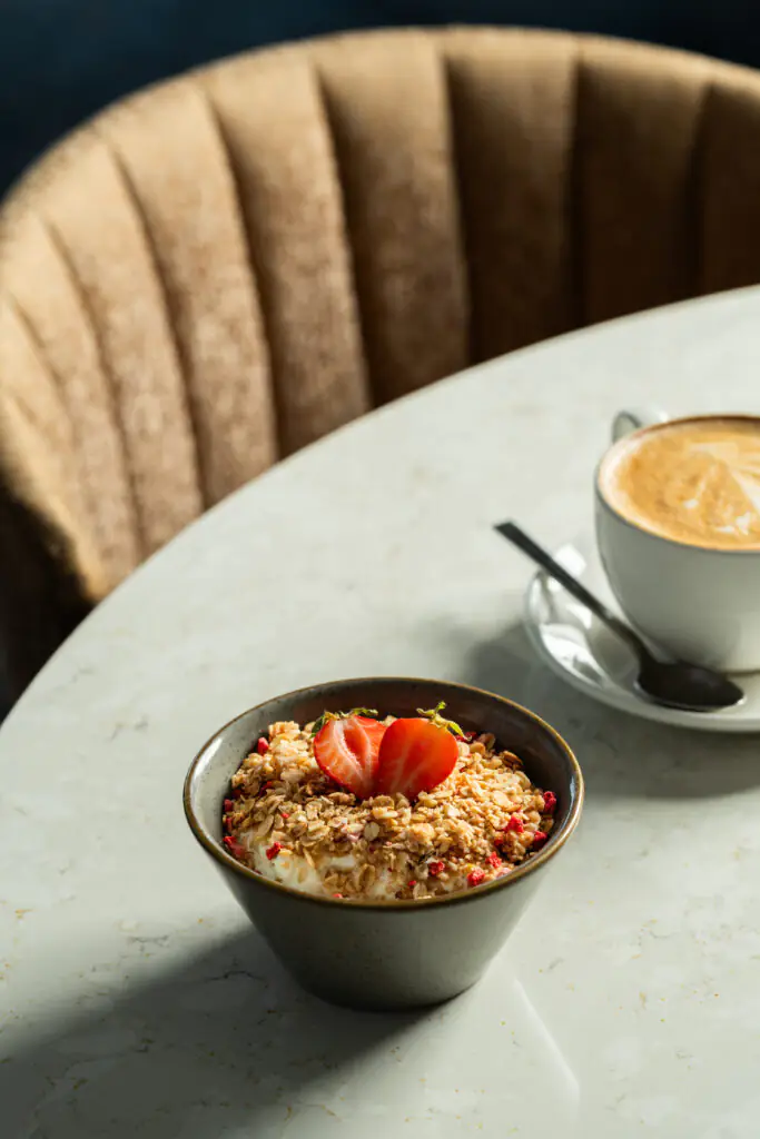 A bowl of yogurt topped with granola and sliced strawberries next to a cup of cappuccino on a round marble table.