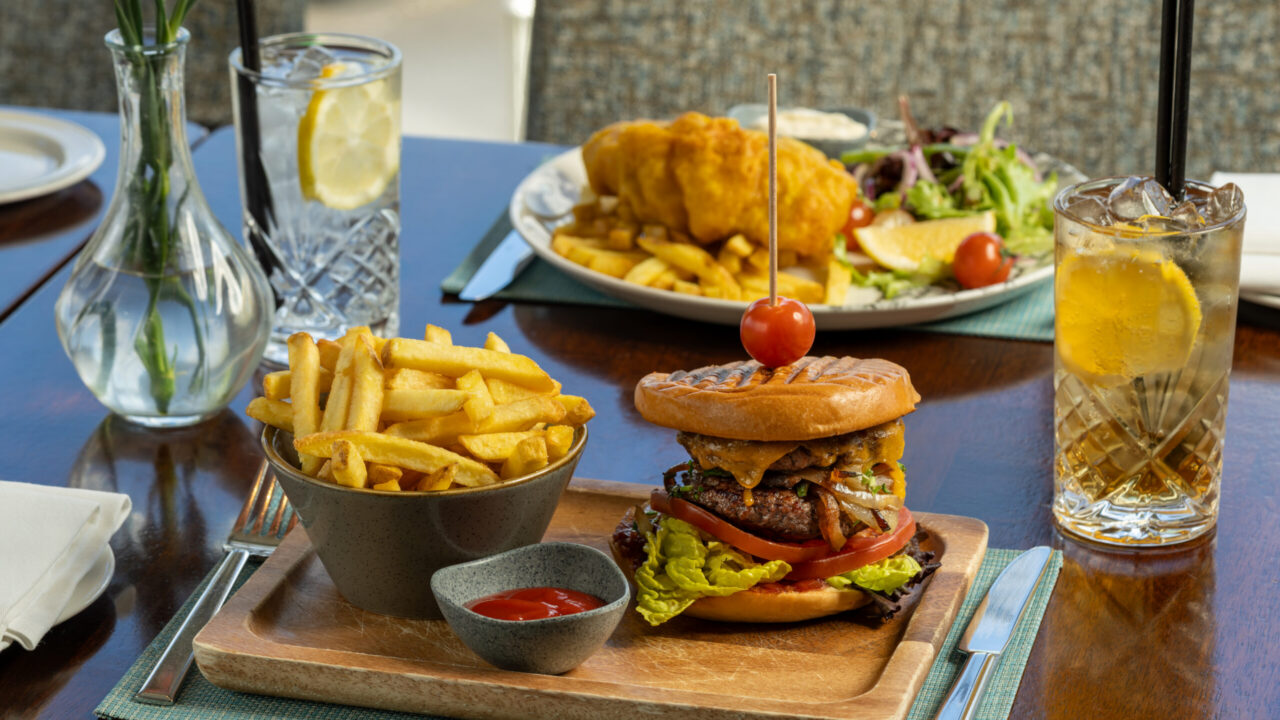 A wooden platter with a burger, fries, and ketchup is on a table next to glasses of iced tea and water. A plate of fish and chips with salad is in the background.