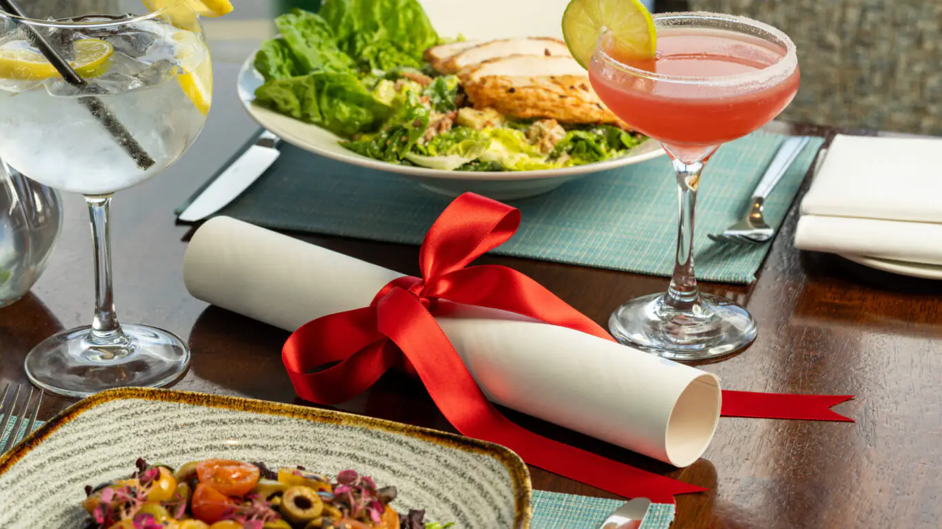 A table set with a salad, pasta, a pink cocktail with a lime, a glass of water with lemon, and a rolled-up scroll tied with a red ribbon.