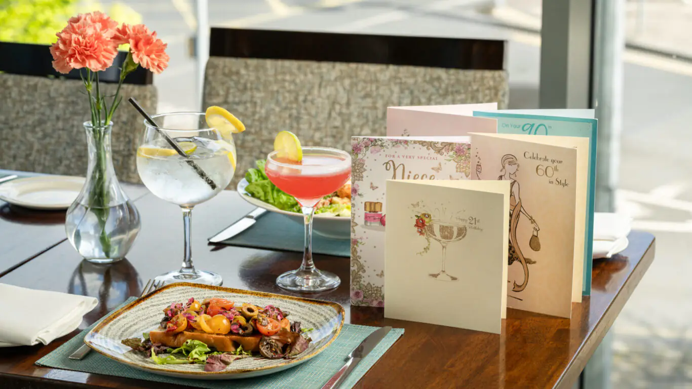 A table with a salad plate, two cocktails, napkins, pink flowers in a vase, and several greeting cards, set near a window overlooking a road.
