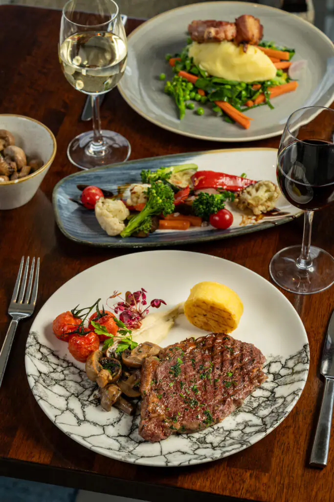 A dinner table with a plate of grilled steak, roasted tomatoes, mushrooms, polenta, and greens, along with wine glasses and additional plated meals in the background.