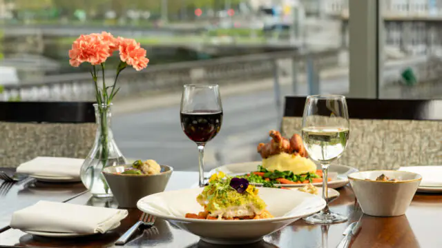A restaurant table set for two with plated meals, bread, two glasses of wine, and a vase of pink flowers, positioned beside a large window overlooking a river.