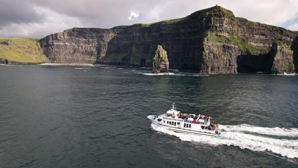 A white sightseeing boat with passengers sails near the Cliffs of Moher, with steep green and rocky cliffs in the background under a cloudy sky.