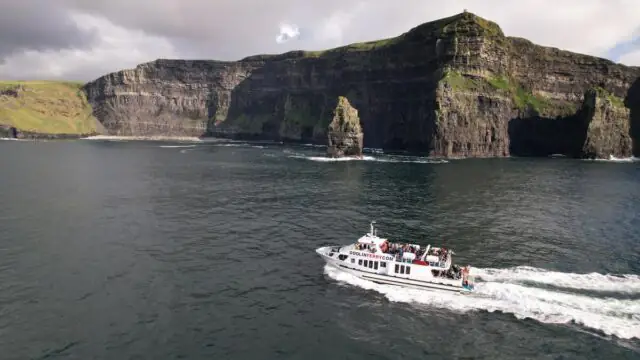 A white sightseeing boat with passengers sails near the Cliffs of Moher, with steep green and rocky cliffs in the background under a cloudy sky.