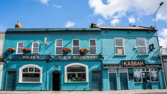 A two-story turquoise building with flower boxes, housing Dolan's Pub & Restaurant and Kasbah Social Club, on a sunny day with a blue sky and clouds.
