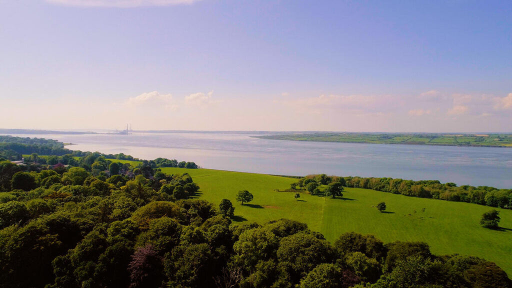 Aerial view of a green field bordered by trees with a wide river or estuary in the background under a clear blue sky.