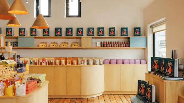 A modern shop interior with wooden shelves and counters displaying books, packaged goods, and jars, under warm lighting and two small windows.