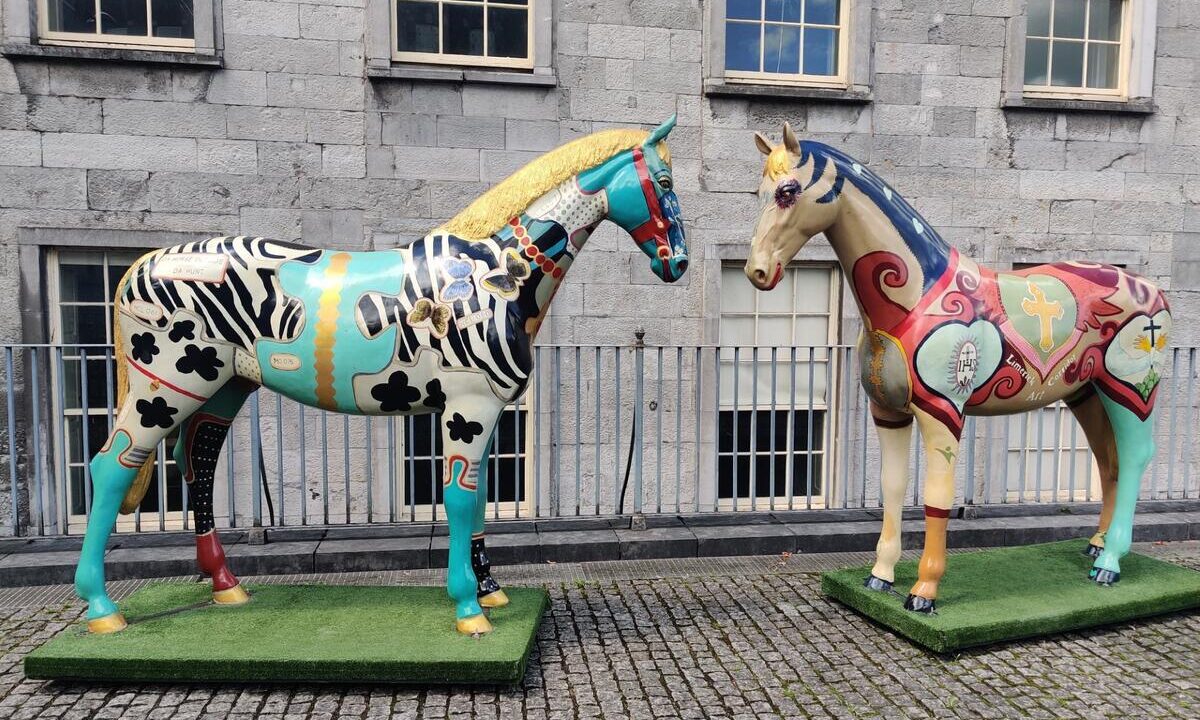 Two colorful, patterned horse statues stand on artificial grass bases in front of a stone building with barred windows.