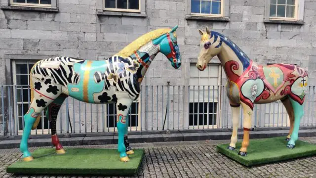Two colorful, patterned horse statues stand on artificial grass bases in front of a stone building with barred windows.