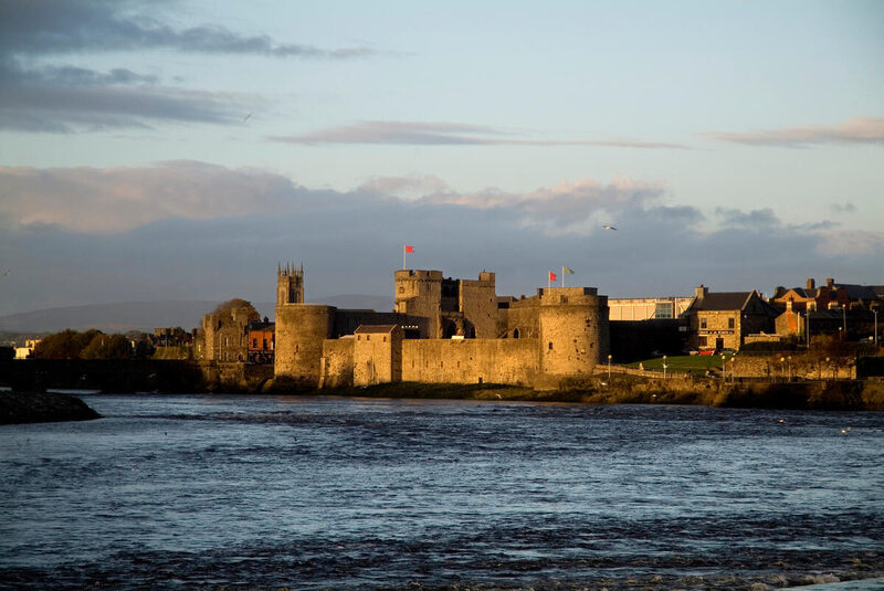 A stone castle with round towers and red flags stands near a river, surrounded by buildings under a partly cloudy sky at sunset.