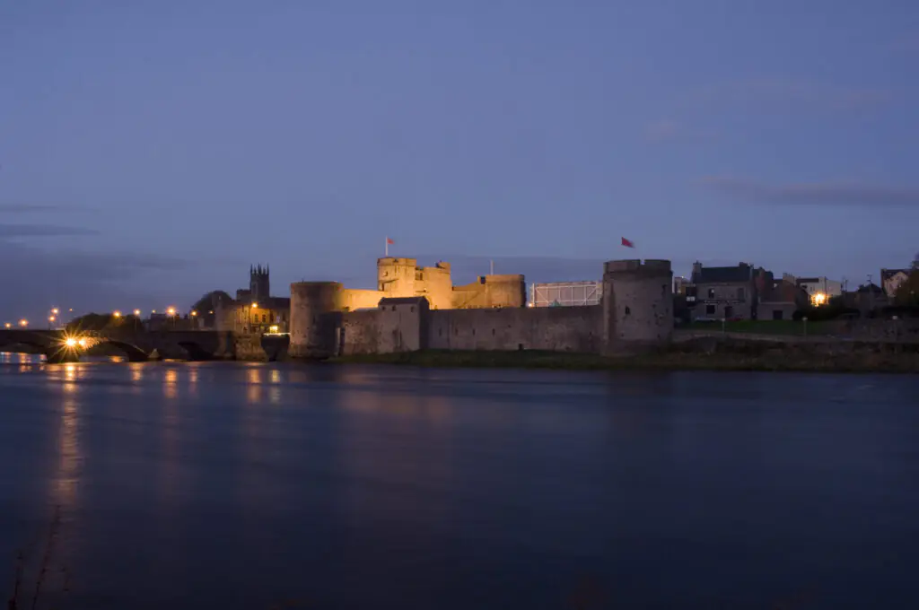 A stone castle with illuminated towers stands beside a river at dusk, with a bridge and buildings visible in the background.