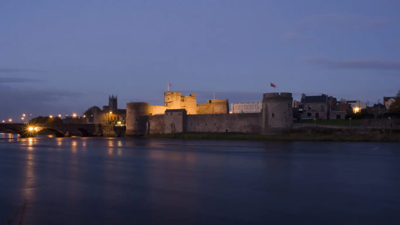 A stone castle with illuminated towers stands beside a river at dusk, with a bridge and buildings visible in the background.