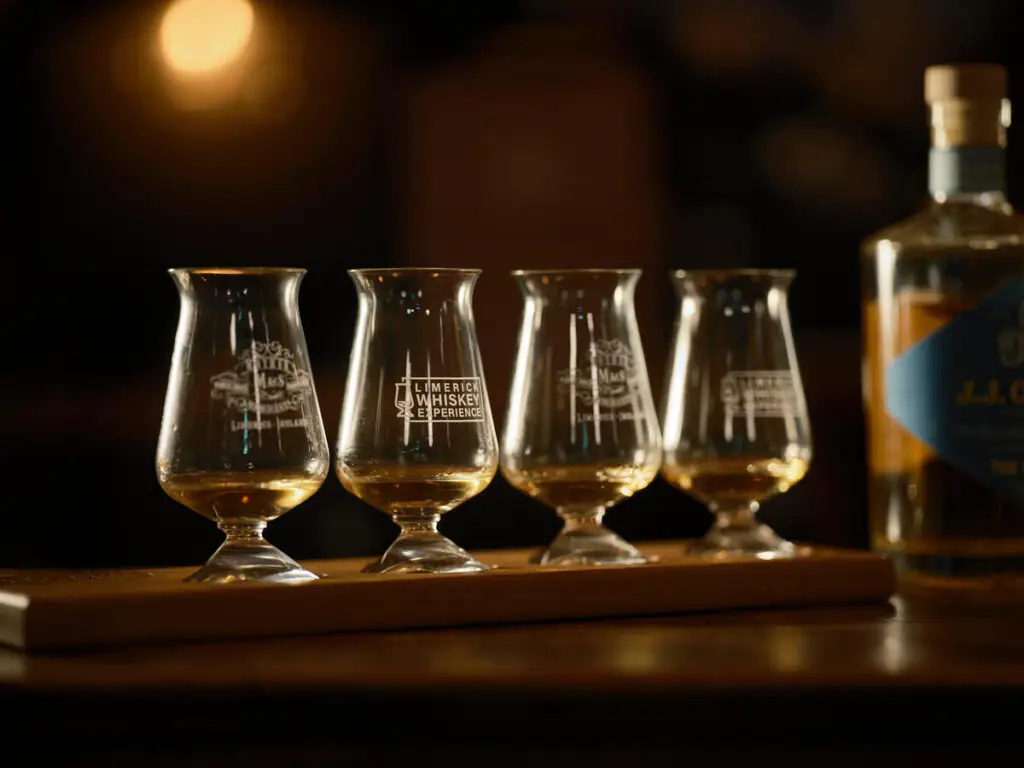 Four tasting glasses with small amounts of whiskey are lined up on a wooden tray; a bottle of whiskey is partially visible to the right.