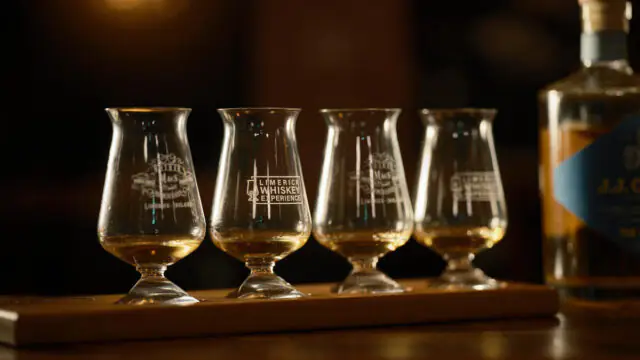 Four tasting glasses with small amounts of whiskey are lined up on a wooden tray; a bottle of whiskey is partially visible to the right.