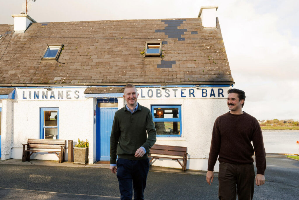 Two men walk outside Linnane’s Lobster Bar, a white stone building with a blue door and benches, under a partly cloudy sky.