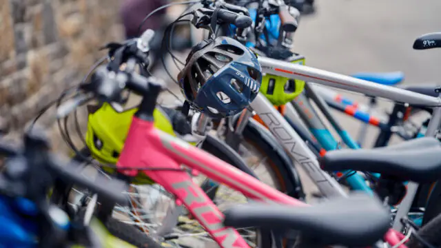 Several colorful bicycles are parked closely together, with helmets hanging from the handlebars, near a stone wall and a paved area.
