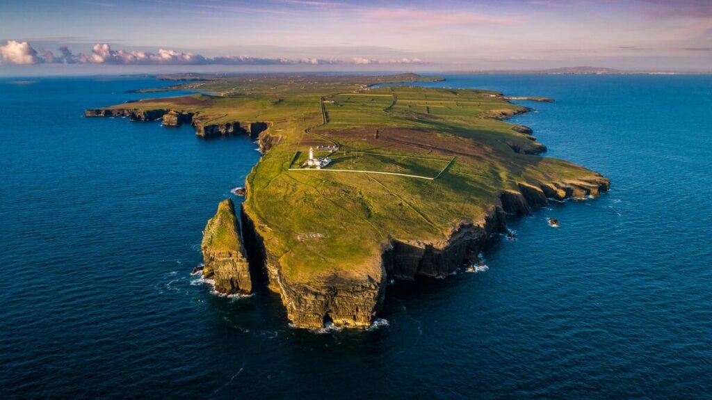 Aerial view of a green, rocky peninsula jutting into the blue ocean with a lighthouse and scattered buildings near the cliff edge under a partly cloudy sky.