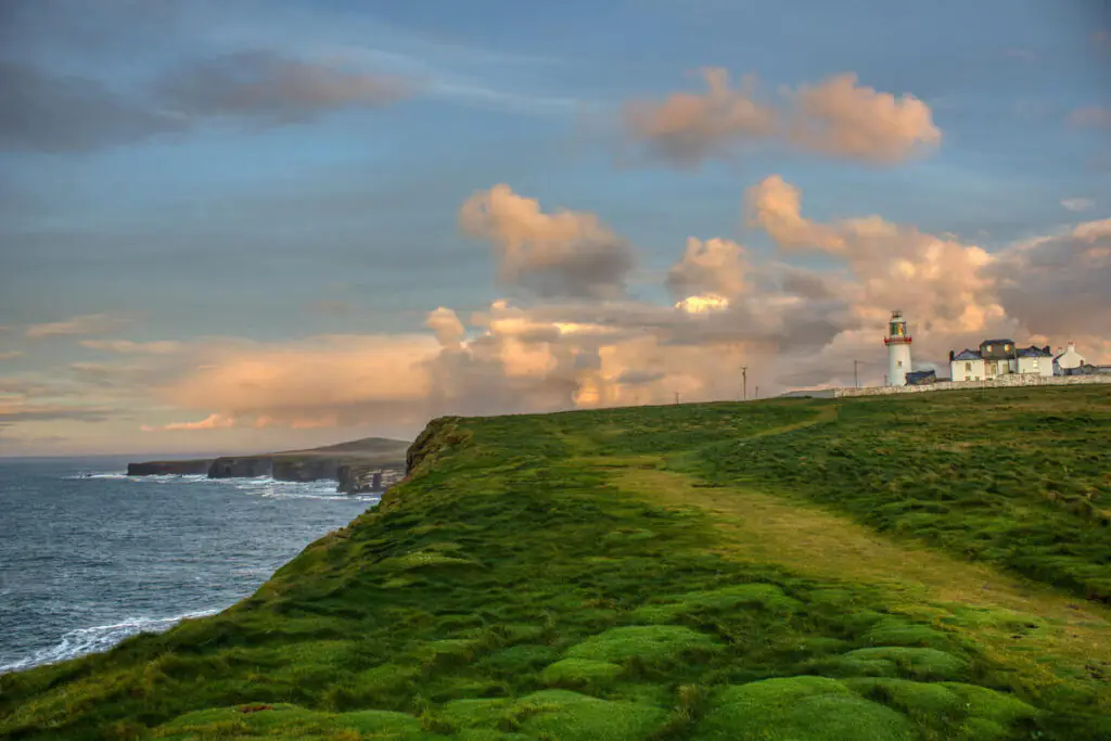 A lighthouse and buildings stand on a grassy cliff overlooking the ocean, with a cloudy sky and rugged coastline in the background.