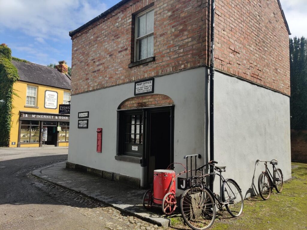 A small brick post office building with a red mailbox, vintage carts, and bicycles outside. Nearby, there is a yellow building and a cobblestone street.