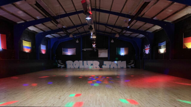 An empty indoor roller skating rink with colored lights on the wooden floor, flags on the walls, and &ldquo;ROLLER JAM&rdquo; painted at the far end.