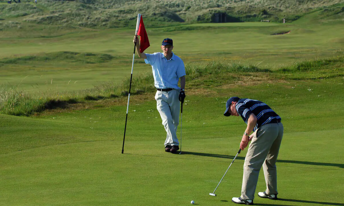 Two men on a golf course; one man is holding the flagstick while the other is putting a golf ball toward the hole on the green.