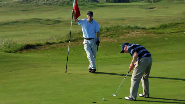 Two men on a golf course; one man is holding the flagstick while the other is putting a golf ball toward the hole on the green.