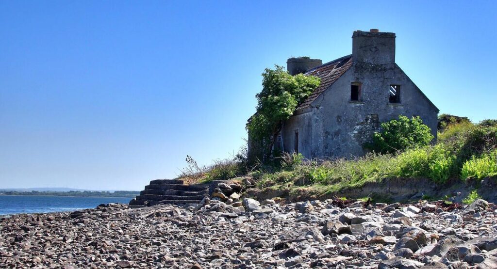 An old, abandoned stone house with a damaged roof sits by a rocky shoreline under a clear blue sky.